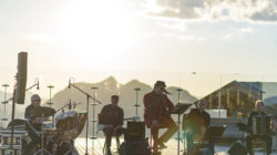 Eine Band spielt bei Sonnenuntergang im Freien mit Schlagzeug, Keyboard, Gitarre und Akkordeon auf einer Holzbühne.