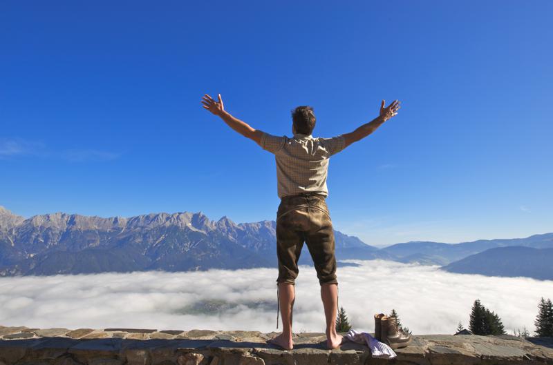 Mann steht barfuß auf einer Steinmauer mit erhobenen Armen und blickt auf Berge über einem Wolkenmeer.