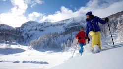 Zwei Schneeschuhwanderer im Tiefschnee mit verschneiten Bergen und Kiefern im Hintergrund unter blauem Himmel.
