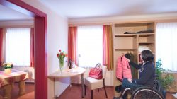 Eine Frau im Rollstuhl hängt eine Jacke in einem barrierefreien Hotelzimmer mit Schreibtisch und offenem Kleiderschrank auf.