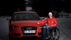 Ein Mann im Rollstuhl in einer roten Jacke posiert neben einem roten Audi auf einer verschneiten Straße.