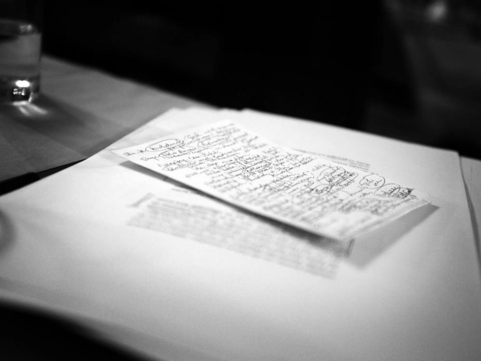 Ein handgeschriebener Zettel mit Kritzeleien und Bearbeitungen liegt auf einem Stapel Papiere auf einem Tisch.