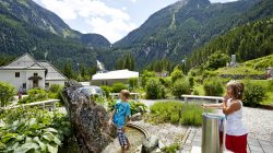 Zwei Kinder spielen an einem Wasserspiel in einem üppigen Berggarten mit Gebäuden und bewaldeten Hügeln im Hintergrund.