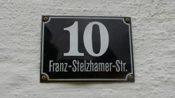 Ein schwarz-weißes Adressschild mit der Nummer 10 Franz-Stelzhamer-Str. auf einer strukturierten weißen Wand.