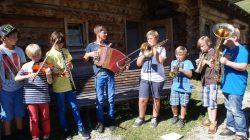 Acht Kinder spielen an einem sonnigen Tag vor einer Holzhütte verschiedene Musikinstrumente.