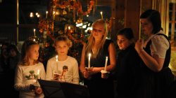 Fünf Kinder stehen mit Kerzen in der Hand vor einem geschmückten Weihnachtsbaum in einem warm beleuchteten Raum.