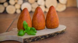 Drei kegelförmige würzige Würste auf einem Holzbrett, mit Basilikumblättern und gestapelten Holzscheiten im Hintergrund.