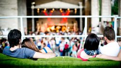 Zwei Paare sitzen auf einer grünen Couch und verfolgen ein Konzert auf einer Freilichtbühne mit einer Menschenmenge darunter.