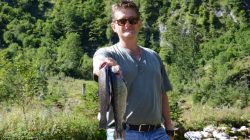 Mann, der zwei große Fische im Freien hält, mit Grünzeug und einem Bach im Hintergrund.