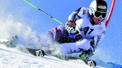 Ein Skifahrer im Rennanzug und mit Helm macht eine scharfe Kurve auf einer verschneiten Piste und wirbelt dabei Schnee auf, während eines Slalomrennens.