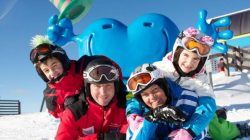 Vier Personen in Skikleidung lächeln, sie liegen im Schnee, hinter ihnen eine blaue Cartoon-Statue und ein klarer Himmel.