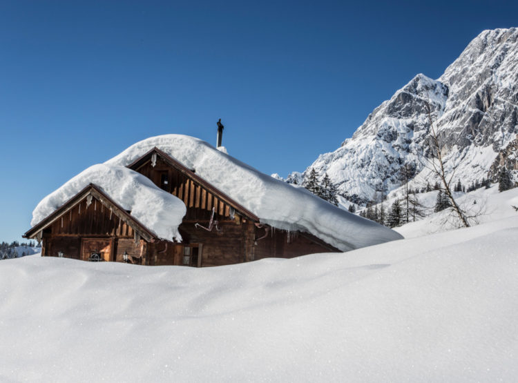 Eine schneebedeckte Holzhütte steht vor verschneiten Bergen unter einem strahlend blauen Himmel.