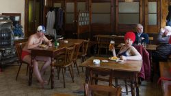 Zwei Personen in Badeanzügen und Mützen essen und trinken in einer Kneipe; im Hintergrund sitzen weitere Gäste, die vollständig bekleidet sind.