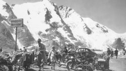 Menschen und Motorräder, die in der Nähe von verschneiten Bergen geparkt sind; ein Mann und ein Kind gehen im Vordergrund, Oldtimer dahinter.