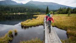 Zwei Radfahrer fahren auf einem schmalen Holzweg über das Wasser mit Bergen und Wald im Hintergrund.