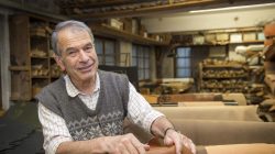 Ein älterer Mann lächelt, während er in einer Werkstatt steht, die mit Leder und Handwerksmaterialien gefüllt ist.