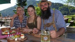 Drei Personen in traditioneller österreichischer Kleidung genießen Essen und Bier an einem Tisch im Freien mit den Bergen im Hintergrund.