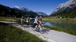 Vier Personen fahren mit dem Fahrrad auf einem Weg neben einem Fluss, mit Bergen und blauem Himmel im Hintergrund.