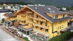 Ein großes gelbes Hotel im alpinen Stil mit Balkonen, Sitzgelegenheiten im Freien und Bergblick im Hintergrund.