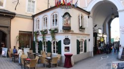 Außencafé mit Tischen und Stühlen neben einem historischen Gebäude mit Fahnen und einem gewölbten Durchgang in einer europäischen Stadt.