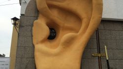 Eine riesige, gelbe Ohrenskulptur ist an der Außenwand eines grauen Gebäudes in der Nähe eines Eingangs angebracht.