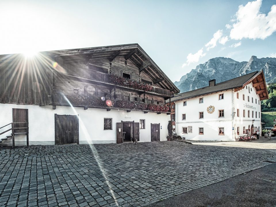 Traditionelle österreichische Alpenhäuser mit Blumenkästen, Sonnenfackeln und Berggipfeln im Hintergrund.