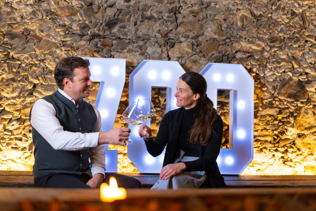 Ein Mann und eine Frau stoßen mit Weingläsern an und sitzen vor großen beleuchteten 700er-Nummern und einer Steinmauer.