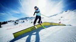 Skifahrer in der Luft bei einem Trick auf einer gelben Schiene in einem verschneiten Geländepark in den Bergen bei strahlendem Sonnenschein.