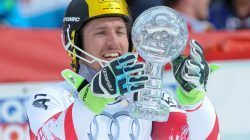 Ein Skifahrer in Rennkleidung hält eine große Kristalltrophäe und lächelt bei einem Skirennen.