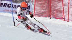 Ein Skifahrer im weißen Anzug und mit orangefarbenem Helm fährt auf einer verschneiten Piste um ein Tor herum.