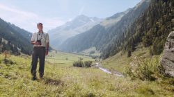 Ein Mann in Försteruniform steht in einem sonnigen, grünen Bergtal mit einem Bach und fernen Gipfeln.
