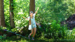 Eine lächelnde Frau balanciert auf einer Slackline in einem sonnenbeschienenen, grünen Wald.