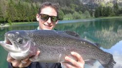 Ein lächelnder Mann mit Sonnenbrille hält einen großen Forellenfisch an einem malerischen See mit Bäumen und Bergen im Hintergrund.