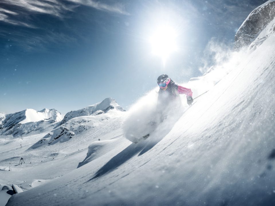 Ein Skifahrer fährt bei strahlendem Sonnenschein und klarem Himmel einen verschneiten Berghang hinunter.