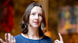 Frau mit schulterlangem Haar gestikuliert mit den Händen und spricht, der Hintergrund ist unscharf.