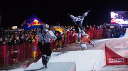 Vier Athleten in Schlittschuhen rennen und springen nachts auf einer verschneiten Bahn, beobachtet von einer jubelnden Menge.