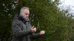 Ein Mann im Freien hält eine Steinschleuder und bereitet sich auf das Zielen vor, im Hintergrund sind Bäume zu sehen.