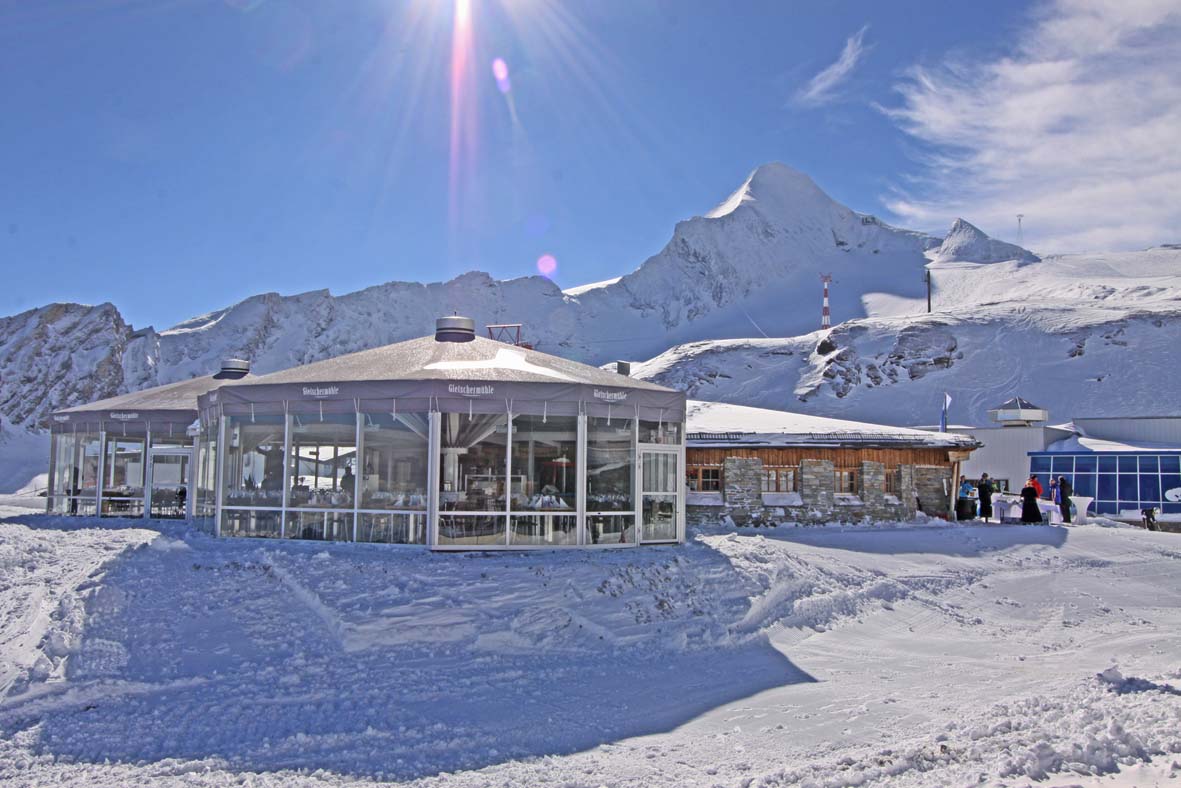 Ein gläsernes Restaurant liegt in einer verschneiten Berglandschaft unter einem hellen, sonnigen Himmel.