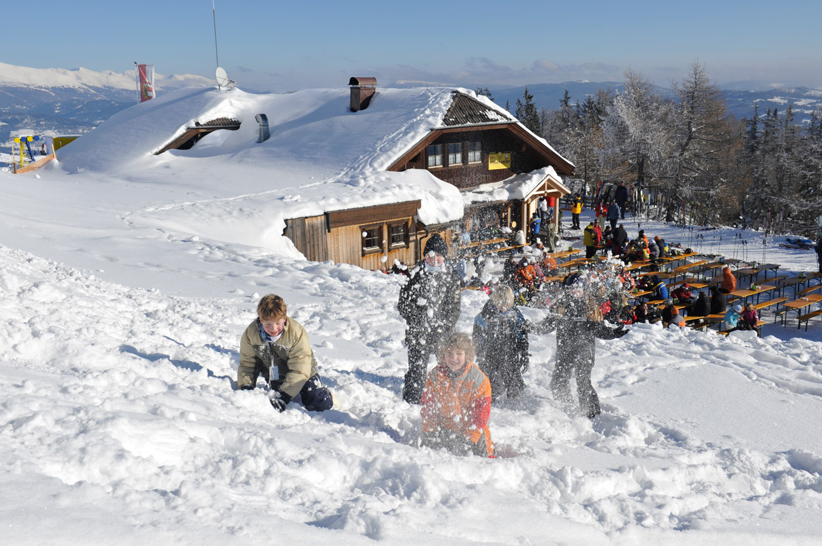 Kinder spielen im Schnee in der Nähe einer Hütte mit Menschen, die draußen an Tischen in einer verschneiten Berglandschaft sitzen.