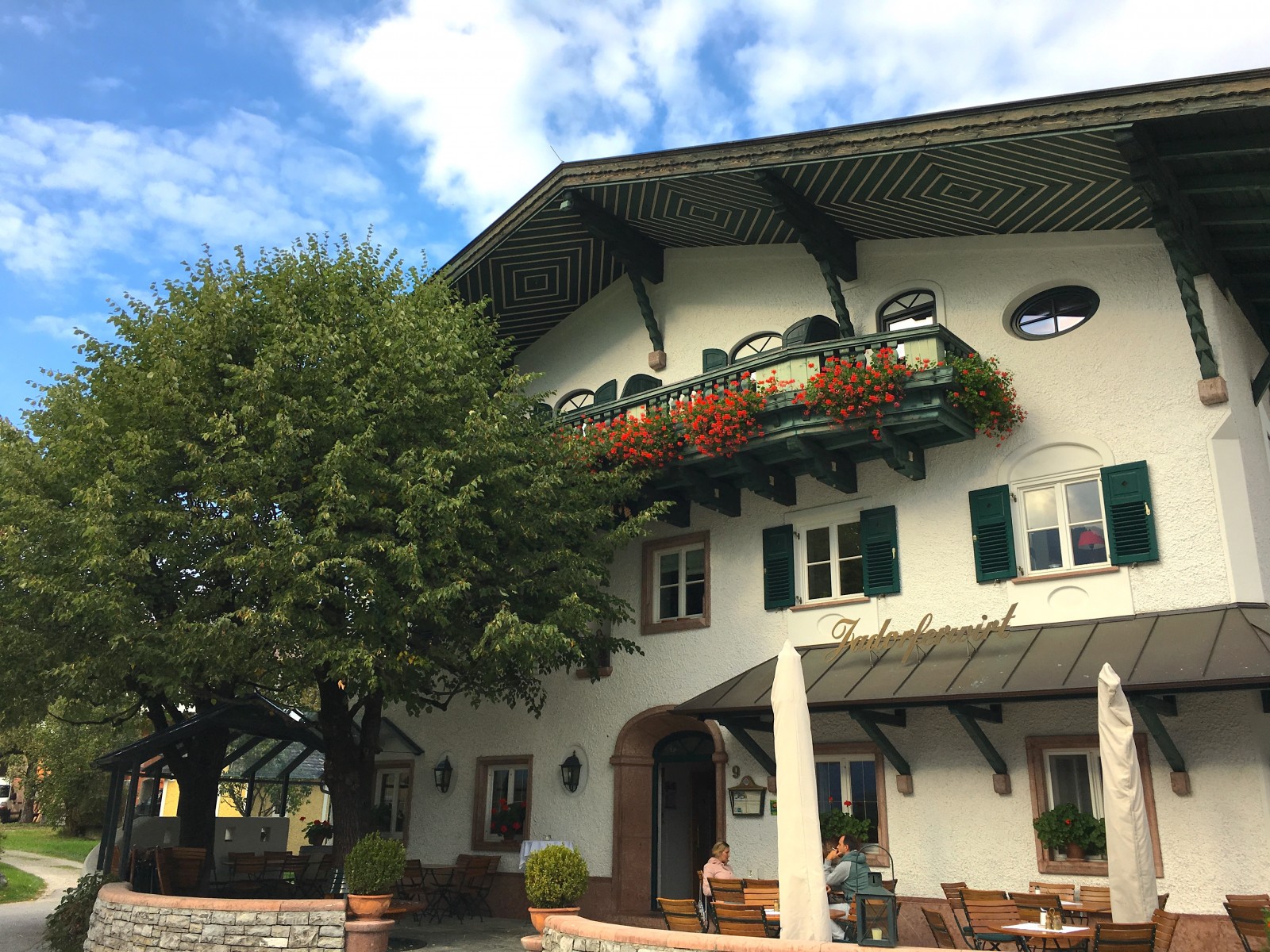 Charmantes Gebäude im alpinen Stil mit grünen Fensterläden, Blumenkästen und Sitzgelegenheiten im Freien unter einem Baum.