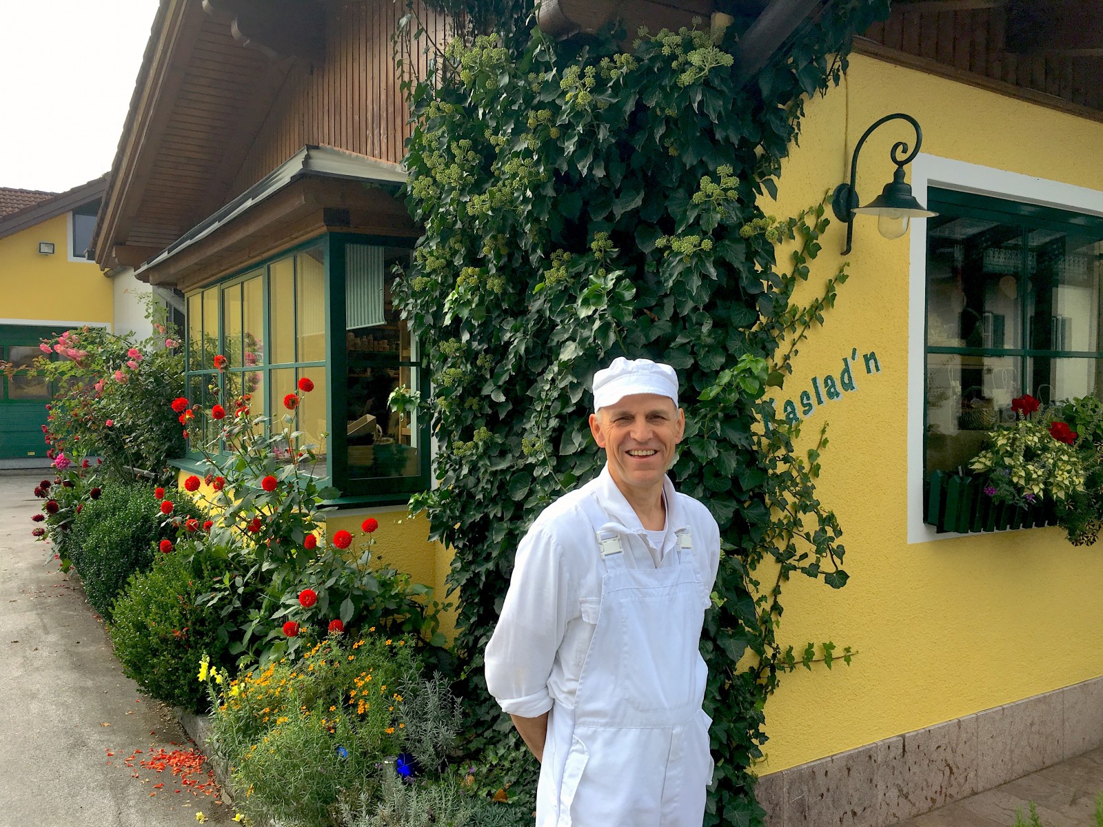 Ein lächelnder Bäcker in weißer Uniform steht neben einem gelben Gebäude mit üppigen Blumen und Grünpflanzen.