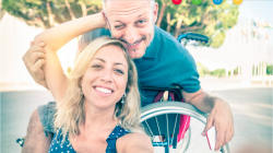 Eine lächelnde Frau und ein Mann im Rollstuhl machen gemeinsam ein fröhliches Selfie im Freien.