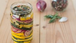 Ein Glas mit eingelegtem gelbem Kürbis, Zucchini und roten Zwiebelscheiben auf einem Holztisch, daneben Kräuter und Gewürze.