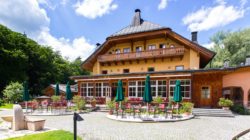 Ein großes Gebäude im Chalet-Stil mit Sitzplätzen im Freien und grünen Sonnenschirmen, umgeben von Bäumen und blauem Himmel.