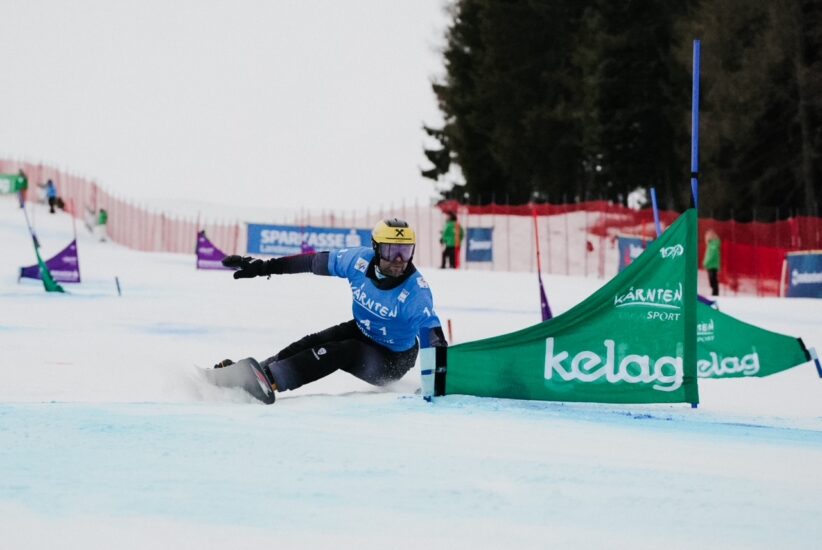 Ein Snowboard Weltmeister aus dem SalzburgerLand in blau rast um eine grüne Flagge auf einem verschneiten Slalomkurs.