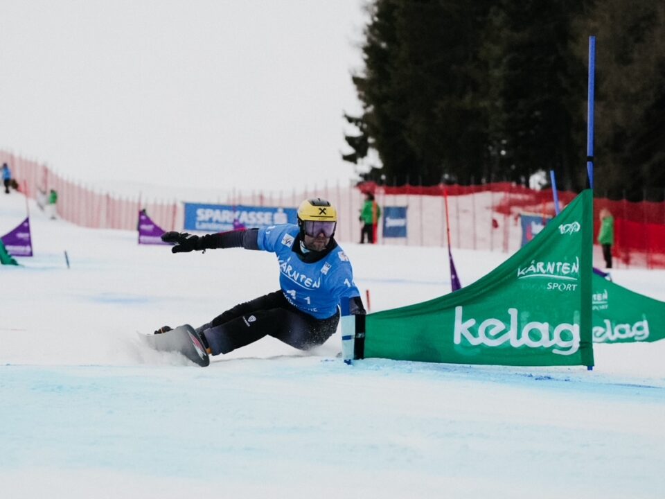 Ein Snowboard Weltmeister aus dem SalzburgerLand in blau rast um eine grüne Flagge auf einem verschneiten Slalomkurs.