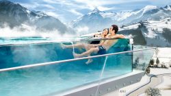 Ein Paar entspannt sich in einem Infinity-Pool im Freien mit Blick auf verschneite Berge unter einem sonnigen Himmel.