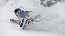 Snowboarder in farbenfrohen Klamotten carven durch den Tiefschnee und erzeugen dabei eine Schneefontäne um sich herum.