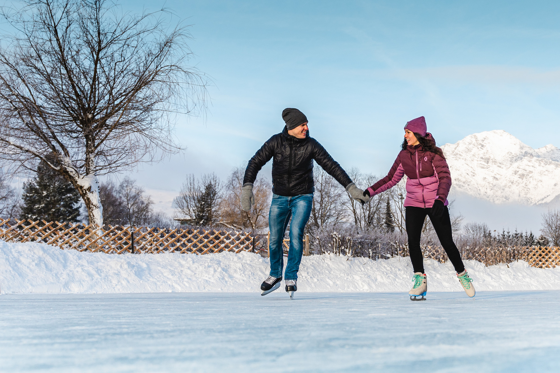 Ein Mann und eine Frau, die im Freien Schlittschuh laufen und sich an den Händen halten, mit verschneiten Bergen und Bäumen im Hintergrund.
