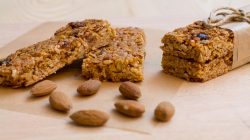 Müsliriegel und ganze Mandeln auf einer Holzunterlage, wobei zwei Riegel mit braunem Papier und Schnur zusammengebunden werden.